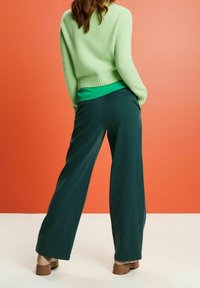 Zelený pletený svetr spárovaný s tyrkysovým topem, širokými tmavě zelenými kalhotami a béžovými botami; hladká textura, volný střih a délka ke kotníkům.