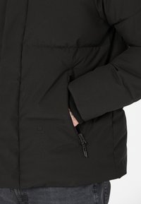 Svart puffjacka med en quiltad design, som har en dragkedjeficka och ribbade ärmslut, tillverkad av strukturerat, vattenavvisande material.