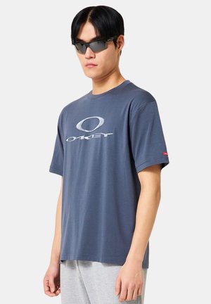 Homme portant des lunettes de soleil foncées, un t-shirt bleu avec le logo Oakley et un pantalon gris clair, debout devant un fond clair uni.
