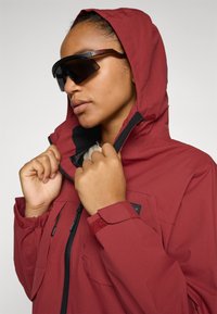 Red waterproof jacket with a hood and zippered front, featuring black accents, adjustable cuffs, and a high collar; paired with dark sunglasses.