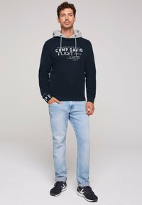 Sudadera navy con capucha gris forrada, que presenta detalles en texto blanco. Combinada con jeans azul claro y zapatillas negras con acentos blancos.