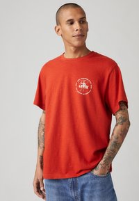 Raudona medvilninė marškinė su apvalia iškirpte, trumpomis rankovėmis ir baltu Levi's logotipu, atspausdintu ant krūtinės. Atsipalaidavęs kirpimas su sklandžiu audiniu.