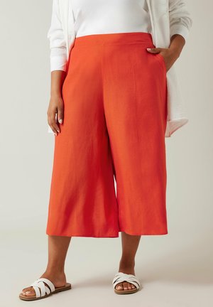 Helle orange Culotte-Hosen mit einer glatten Textur, die Seitentaschen und eine lockere Passform aufweisen, kombiniert mit weißen Sandalen mit Überkreuzriemen.