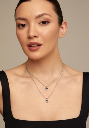 Mujer con cabello oscuro y top negro sin mangas, luciendo dos collares plateados con colgantes de piedras preciosas azules sobre un fondo beige.