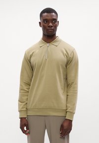 Khaki színű, hosszú ujjú póló pulóver cipzáras gallérral, puha anyagból készült. Fitting szabású, bordás mandzsettákkal és aljjal rendelkezik.