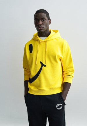 Jeune homme portant un sweat à capuche jaune vif avec un grand motif smiley noir, associé à un pantalon de survêtement noir Moschino, debout devant un fond uni.