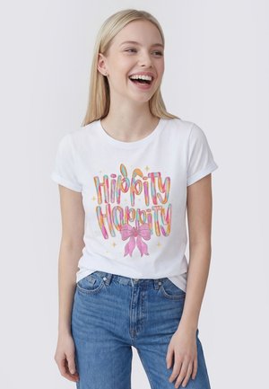 Jeune femme souriante portant un t-shirt blanc avec le texte coloré « Hippity Hoppity » et un nœud rose, associé à un jean bleu.