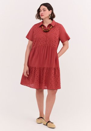 Frau trägt ein kurzärmeliges rostrot Kleid mit Lochstickereidetails, eine auffällige Halskette, Creolen und beige geflochtene flache Schuhe.