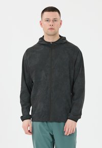 Schwarze Zip-Hoodiejacke mit einem dezenten strukturierten Muster, ausgestattet mit elastischen Bündchen und einem lässigen Schnitt. Über türkisfarbenen Hosen getragen.