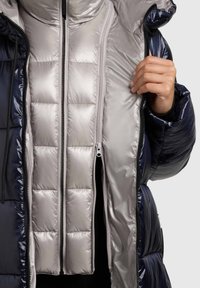 Tollkabát tengerészkék külsővel és ezüst belső béléssel, steppelt részekkel, cipzárokkal és magas gallérral. Puha, fényes textúrák.