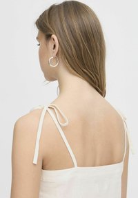 Top de lino blanco con tirantes finos atados en los hombros. Textura suave y diseño sencillo. La modelo lleva pendientes de aro plateados. 