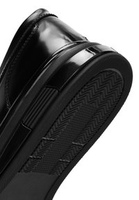 Chaussure habillée noire brillante inclinée pour montrer la semelle en caoutchouc texturée avec des motifs en zigzag et en croix.