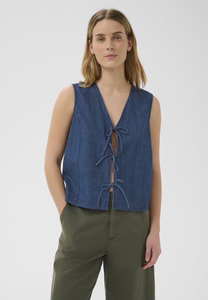 Femme aux cheveux blonds mi-longs porte un top en jean bleu sans manches noué avec trois nœuds et un pantalon vert olive sur fond uni.