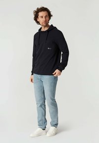 Sudadera navy con mangas largas, un bolsillo frontal y cordones. Combinada con jeans azul claro y zapatillas blancas. Fondo liso.