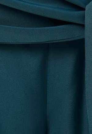 Lähikuva sinivihreän kankaan laskoksista ja vyölenkin yksityiskohdasta vaatteessa, korostaen sileää tekstuuria ja sauman linjoja.