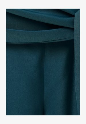 Primer plano de pliegues de tela verde azulado y un detalle de presilla de cinturón en una prenda, destacando la textura suave y las líneas de las costuras.