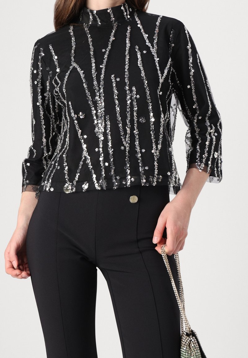Haut noir transparent à manches longues avec des rayures verticales en sequins argentés, associé à un pantalon noir ajusté, tenant un petit sac à bandoulière chaîne.