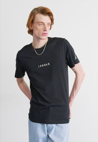 Juoda medvilnės marškinėliai su užrašu "JORDAN" baltu spalva priekyje ir mažuoju logotipu ant kairiojo rankovės. Apykaklė suapvalinta ir trumpomis rankovėmis.