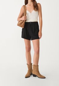 Femme portant un débardeur blanc sans manches, un short noir, des bottes de cow-boy en suède beige, et portant un sac à épaule en suède beige assorti.