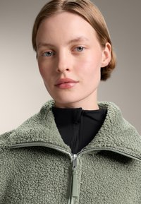 Groene, pluche, ritsjack met een zachte textuur, voorzien van een hoge kraag en een contrasterende zwarte binnenlaag en rits.