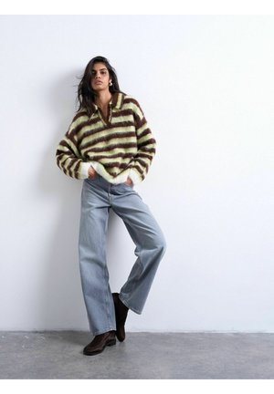 Donna con un maglione felpato a righe verdi e marroni, jeans a gamba larga azzurri chiari e stivaletti marrone scuro, in piedi contro un muro bianco.