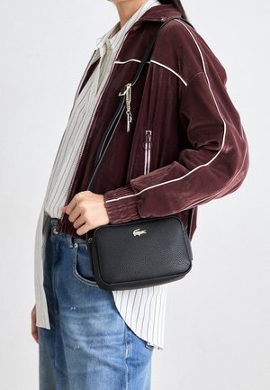 Personne portant une veste bordeaux zippée, une chemise blanche à rayures, un jean bleu, portant un petit sac bandoulière noir texturé avec un logo argenté.
