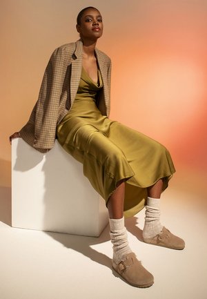 Khaki satin dress with a cowl neckline, paired with a plaid blazer. Beige suede shoes and knitted cream socks. Model seated on a white pedestal.