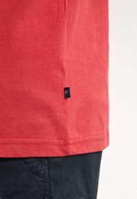 Camiseta de algodón roja con una textura suave, que presenta una pequeña etiqueta azul marina en el dobladillo. La parte inferior tiene un corte recto y detalles de costura visibles.