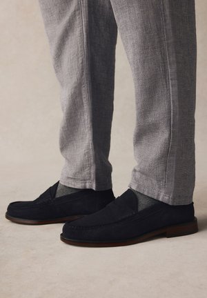 Gambe con pantaloni grigio chiaro strutturati, calzini grigio scuro e mocassini in camoscio nero con suole marroni che stanno su un pavimento neutro.