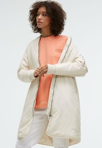 Long, cream quilted coat with a zipper and side pockets, layered over an orange sweatshirt. Text on the sweatshirt reads "SENSES THE LAB."
