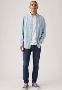 Ανοιχτό μπλε πουκάμισο με κουμπιά πάνω από ένα λευκό T-shirt, συνδυασμένο με σκουρόχρωμα τζιν και μπλε αθλητικά παπούτσια. Το πουκάμισο έχει άνετη γραμμή και μαλακή υφή.