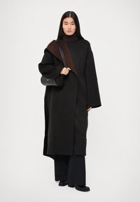 Largo abrigo negro con acento marrón caído sobre el hombro, suéter de cuello alto acanalado, pantalones negros y un bolso negro texturizado.