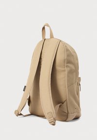 Μπεζ υφασμάτινο σακίδιο πλάτης με επενδεδυμένους ιμάντες ώμου, θήκες με φερμουάρ και λαβή στην κορυφή για εύκολη μεταφορά. Επιφάνεια με υφή και ραφές.