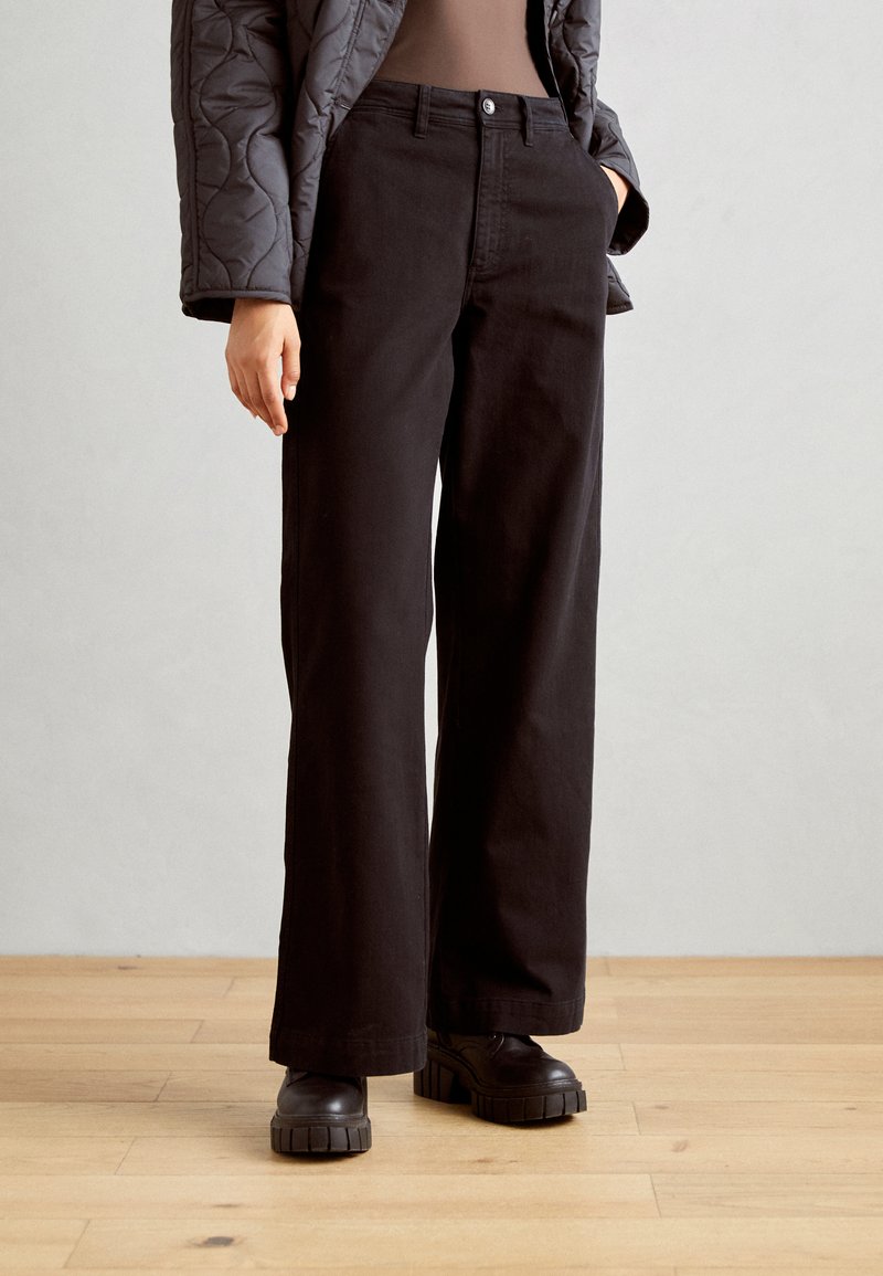 Črne široke hlače iz trdnega bombaža, z gumbom spredaj in stranskimi žepi, v kombinaciji z debelimi črnimi platform čevlji.