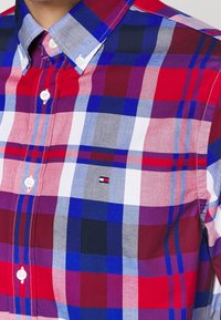 Camisa de cuadros en rojo, azul y blanco con cuello abotonado. Presenta colores contrastantes y un pequeño logo cerca del dobladillo. Hecha de tela texturizada.