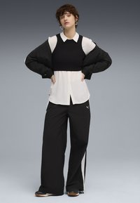 Zwarte gecropte sweater over een wit overhemd met kraag, gecombineerd met zwarte wijde broek met witte zijstrepen en sportieve schoenen.