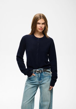 Jeune femme aux cheveux lisses châtain clair portant un cardigan noir boutonné et un jean bleu clair avec une ceinture noire, debout avec une main dans la poche.