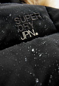 Črna nepremočljiva jakna z vodnimi kapljicami na površini, ki ima izšito logo "SUPER DRY JPN." v kontrastnih barvah.