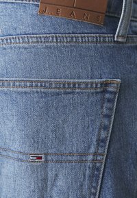 Blå denimjeans med en brun lærlabel merket "JEANS." Funksjoner inkluderer en baklomme med rød og hvit sømdetalj.