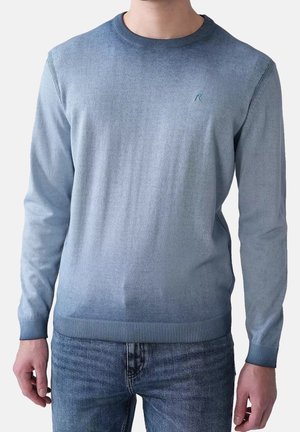 Homme portant un pull à col rond bleu clair et un jean bleu, debout devant un fond uni, les mains détendues le long du corps.