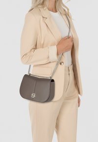 Borsa a spalla in pelle marrone con pattina curva, tracolla in catena argentata e dettaglio del logo, esposta su uno sfondo neutro.