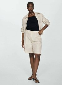 Beige linnen shorts och matchande kort jacka över en svart topp. Modellen poserar med ena handen på höften, iförd svarta sandaler. Slät, strukturerad tyg.