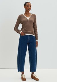 Maglietta marrone a maniche lunghe con colletto bianco e bottoni, abbinata a jeans larghi blu e sneakers marroni con lacci bianchi.