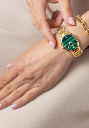 Orologio color oro con quadrante verde decorato, cassa rotonda e bracciale metallico. Una mano regola delicatamente l'orologio mentre poggia su una superficie chiara.