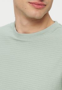 Κοντινό πλάνο στο κάτω μέρος του προσώπου και του λαιμού ενός ατόμου που φοράει ανοιχτοπράσινο ριπ πλεκτό πουλόβερ με στρογγυλή λαιμόκοψη, σε απλό φόντο.