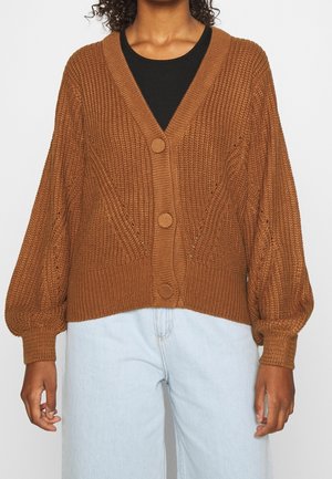 Gilet en tricot marron avec un col en V, des boutons à l'avant et des manches ballon. Présente des motifs texturés et des poignets côtelés, porté par-dessus un haut noir.