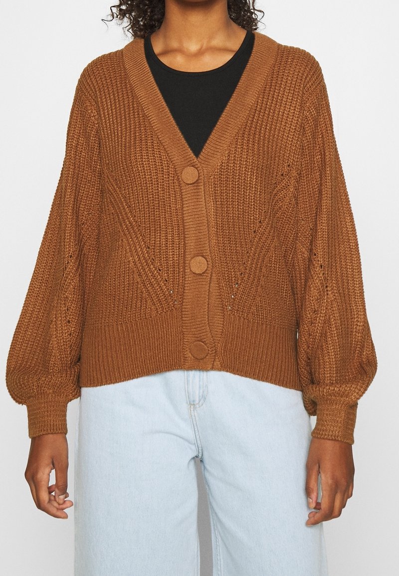 Brun stickad kofta med V-ringning, framknappar och ballongärmar. Har texturerade mönster och ribbade manschetter, bärs över en svart topp.