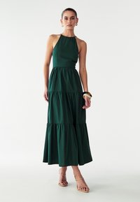 Temno zelena, večslojna maksi obleka iz lahkega materiala, z obliko halter vratu in nabranim zgornjim delom. Kombinirana s nude sandali.