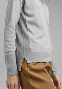 Světle šedý kardigan s výstřihem do V, zapínáním na knoflíky vepředu a žebrovanou texturou po stranách, má boční rozparky a dlouhé rukávy.