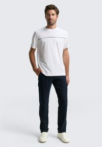 Mann steht mit Händen in den Taschen, trägt ein weißes Tom Tailor T-Shirt, dunkle Cargo-Hosen und weiße Sneakers vor einem einfarbigen Hintergrund.